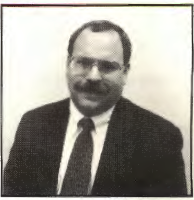 A man with glasses, a mustache, and short hair, wearing a suit, dress shirt, and tie, poses in front of a plain background—representing the professionalism of the CPA Team.